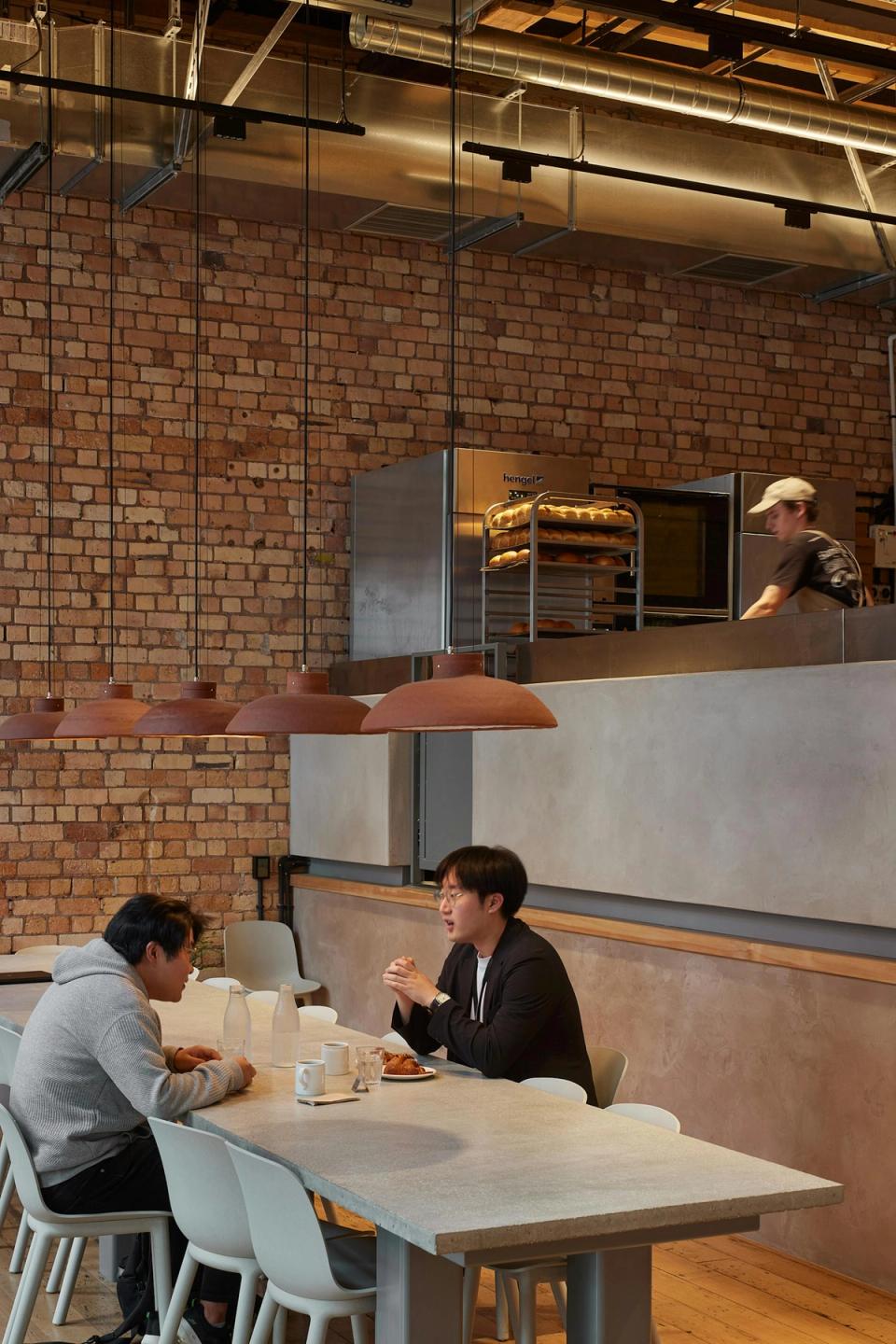Communal dining table poured in situ from local aggregates at Daily Bread Britomart, with hand-built ceramic pendants by Keegan Davis suspended overhead and the open-plan bakery beyond