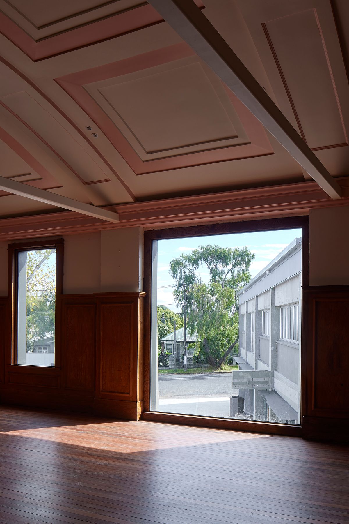Restored heritage interior at The Chambers showing original coffered ceiling and timber wainscoting paired with a large new picture window inserted into the existing facade, opening to the Mount Albert streetscape — adaptive reuse of the 1927 Council building by Knight Associates