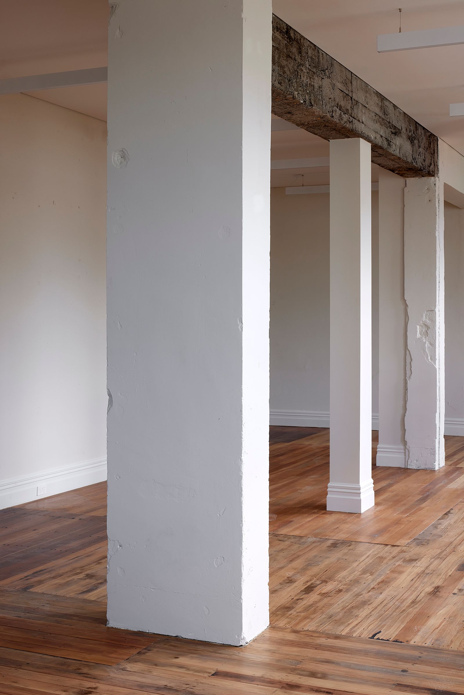 Restored open-plan interior at The Chambers showing structural columns paired with an exposed original concrete beam overhead, set against sanded native timber flooring — adaptive reuse of the 1927 Mount Albert Council building by Knight Associates