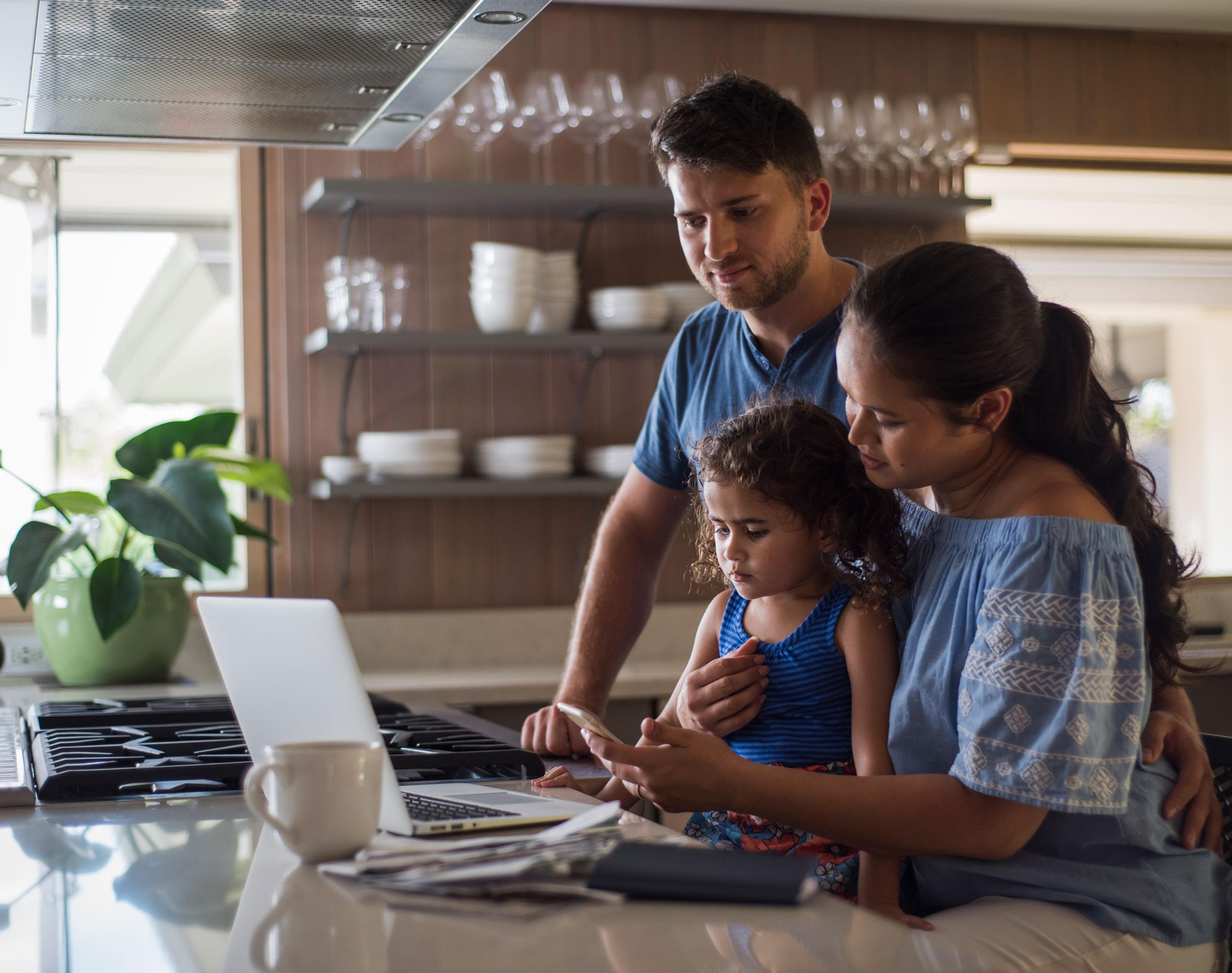 Father, mother, and daughter sitting at kitchen bar looking at a phone and laptop.