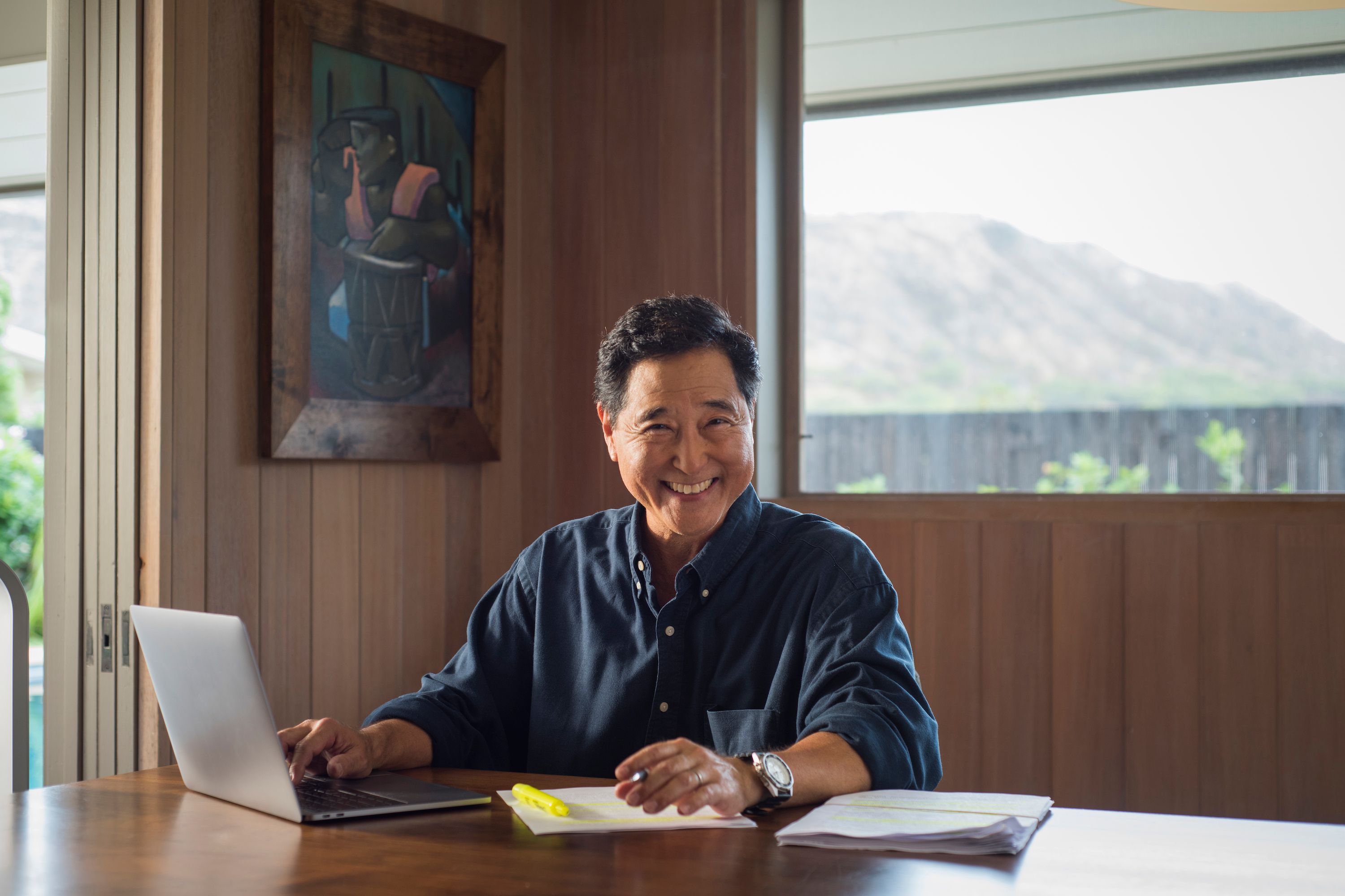 Man with a pen in hand smiling at the camera, seated at a table with various documents.