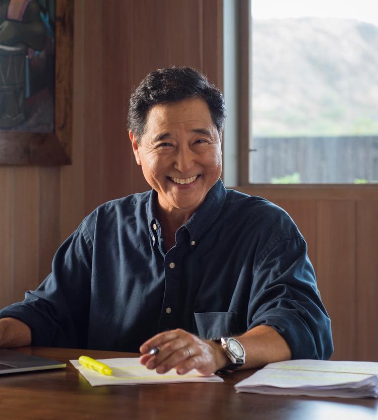 Man with a pen in hand smiling at the camera, seated at a table with various documents.