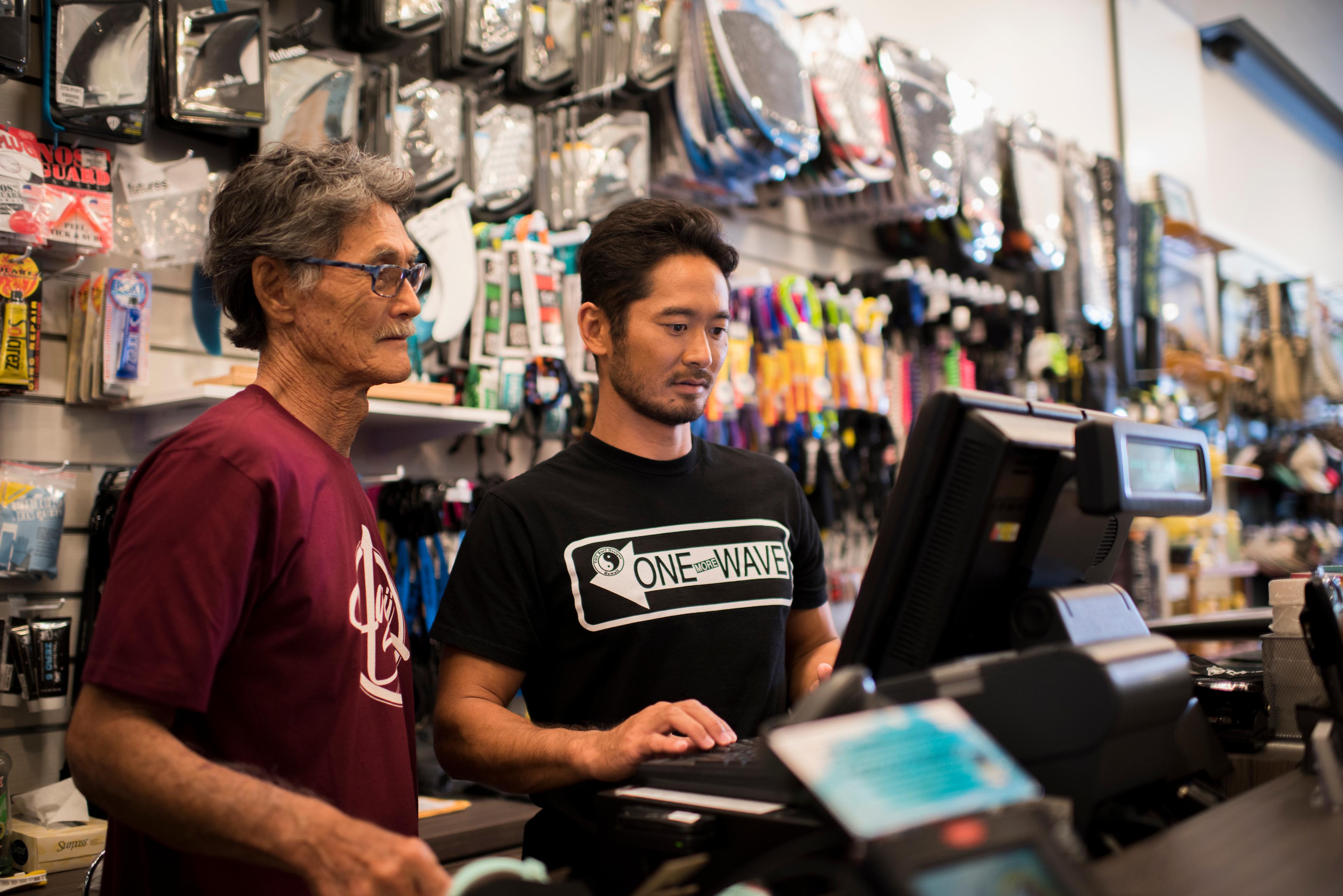 Video cover image shows entrepreneurs Craig and Ryan Sugihara looking at a computer screen inside their store, T&C Surf