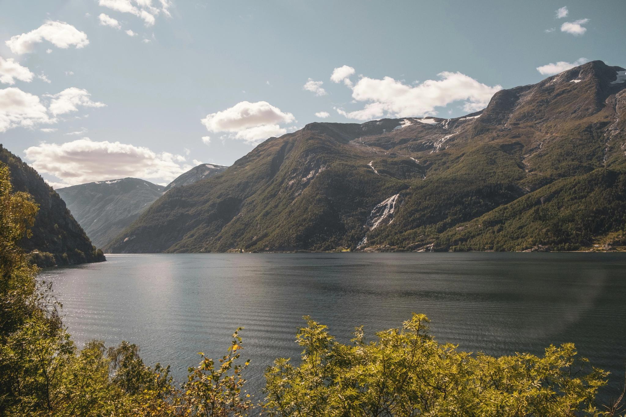 A scenic fjord landscape with steep, forested mountains, some snow and waterfalls, under a partly cloudy sky.