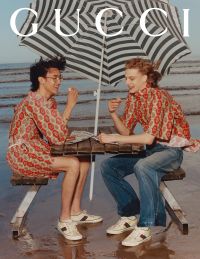 Two adults wearing matching patterned tops and white sneakers sit at a picnic table under a black and white striped umbrella on a beach while eating.