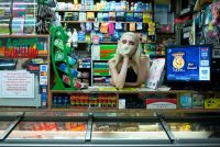 A person with long light hair wearing a sleeveless top is leaning on a counter inside a store filled with various packaged goods and a digital screen displaying a promotional advertisement.