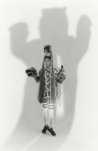 A person with extremely long hair styled upwards and wearing a patterned outfit stands in front of a wall casting a large shadow resembling a bear with raised arms.