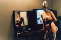 A person wearing a patterned bodysuit with cutouts is leaning on a table in a room with a mirror and a television in the background.