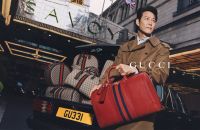 A person wearing a trench coat stands next to a car loaded with Gucci bags outside a building with a Savoy sign.