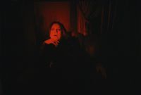 A person with long hair wearing dark clothing and a cross necklace is sitting in a dimly lit room with a surprised or intense expression.