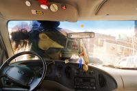 A person with long hair is leaning backward outside a vehicle, viewed through the windshield from inside the car's dashboard and steering wheel area.