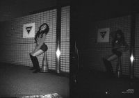 A person with long hair wearing a long-sleeve top, shorts, and knee-high boots is posing against a tiled wall near a traffic cone in a dimly lit urban setting.