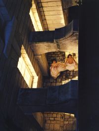 Two people wearing flowing white garments stand on a stone-paved area between illuminated stone walls and arches, viewed from above.