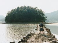 A person wearing a long coat and red socks stands on a rocky path extending into a body of water with a densely forested island in the background.