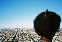 A person with a large afro hairstyle secured with a clip is looking out over a sprawling cityscape under a clear blue sky.