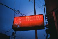A glowing red neon sign reading "DELIRIUM" with a martini glass icon is hanging outdoors against a dark blue evening sky with visible power lines.