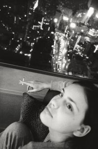 A person with short hair is sitting indoors near a window at night, holding a cigarette and looking up with a calm expression while city lights glow outside.