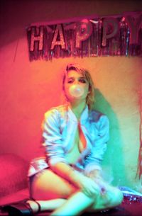 A person with short hair wearing a light-colored jacket and shorts is sitting on the floor blowing a bubble with gum under a "HAPPY" banner in a dimly lit room with colorful lighting.