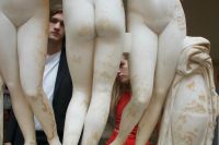 Two people stand behind a row of large, weathered classical statues showing the lower halves of human figures.