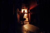 A person is crouching in front of an open refrigerator in a dimly lit room with warm light illuminating the scene.