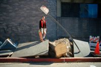 A person wearing a red and black jacket stands on a broken concrete slab holding a skateboard next to a fallen tree trunk and a bent street sign on a city sidewalk.