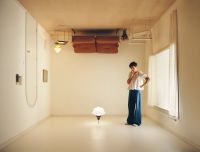 A person wearing a white top and blue pants stands thoughtfully in a minimalistic room with a couch and chairs mounted upside down on the ceiling and a lamp on the floor.