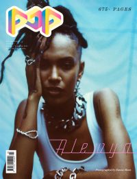 A person with braided hair styled up is wearing a white sleeveless top and chunky silver jewelry while resting their head on one hand against a blue background with colorful text and a barcode on the left side.