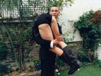 A person dressed in a black suit is holding another person wearing black shorts and black boots in an outdoor garden setting with greenery and a brick wall.