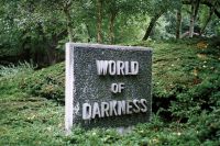 A stone sign with the words "WORLD OF DARKNESS" stands surrounded by green foliage and trees in a natural outdoor setting.