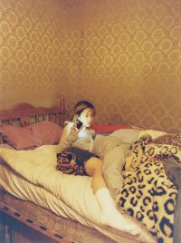 A girl with long hair is sitting on a bed with patterned blankets and pillows, holding a phone and looking towards the camera against a gold patterned wallpaper background.