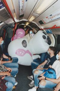 People wearing casual clothes and face masks sit inside a subway car with a girl wearing a large inflatable white cat figure occupying several seats.