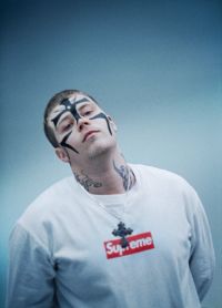 Person with short hair and black face paint wearing a white sweatshirt with a red and white Supreme logo and a large cross necklace against a plain blue background.