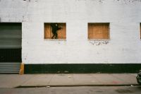 A person dressed in black is climbing through a boarded-up window on a white brick building with a black base and a closed metal garage door.