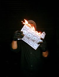 A person wearing black gloves and a dark shirt is holding and burning a sheet of paper with multiple printed dollar bills against a dark background.
