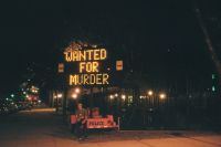 A person is sitting on a bench near a police barricade under a large illuminated sign that reads "WANTED FOR MURDER" on a dimly lit street at night.