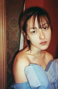 A person with straight hair and bangs is wearing an off-the-shoulder blue and white checkered garment and looking directly at the camera with a neutral expression against a patterned and plain background.