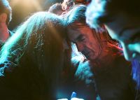 Two people with long hair are closely leaning their heads together in a dimly lit setting with colorful lighting.
