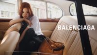 A person with long hair wearing a white shirt and black pants is sitting in the backseat of a vintage car with a brown leather bag beside them and the word BALENCIAGA displayed on the seat.