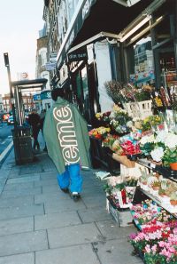 A person wearing a green jacket with large white text on the back walks past a flower shop with colorful bouquets and plants displayed outside on a city sidewalk.