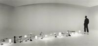 A person stands in a large, empty, curved white space observing a row of various illuminated lamps and light fixtures arranged on the floor.