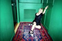 A person with light-colored hair wearing a black dress and sandals is sitting on a patterned rug in a narrow green-walled room while reaching up with one arm.