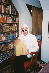 A person wearing white clothing and sunglasses is sitting in a chair holding a wooden box in a room filled with shelves of books and magazines.