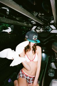 A person wearing a plaid skirt, a white bikini top, and a baseball cap is sitting inside a vehicle with several white birds perched nearby and one bird flying close to the person.