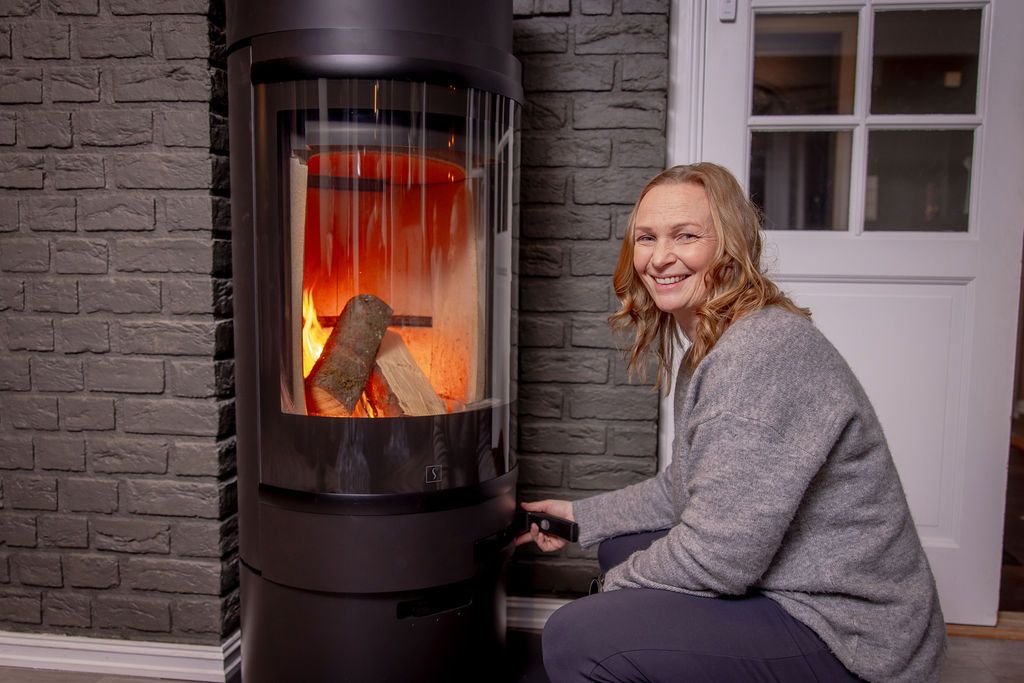Person sitter ved frittstående vedovn og justerer lufttilførsel, med flammer synlige bak glassdør
