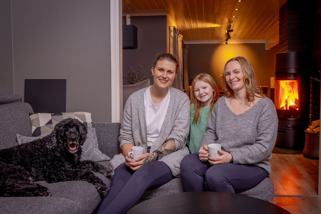 Personer og hund sitter i sofa i stue med frittstående vedovn og synlige flammer i bakgrunnen