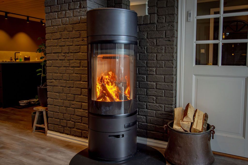 Frittstående rentbrennende vedovn med synlige flammer, plassert mot murvegg, med vedkurv ved siden av