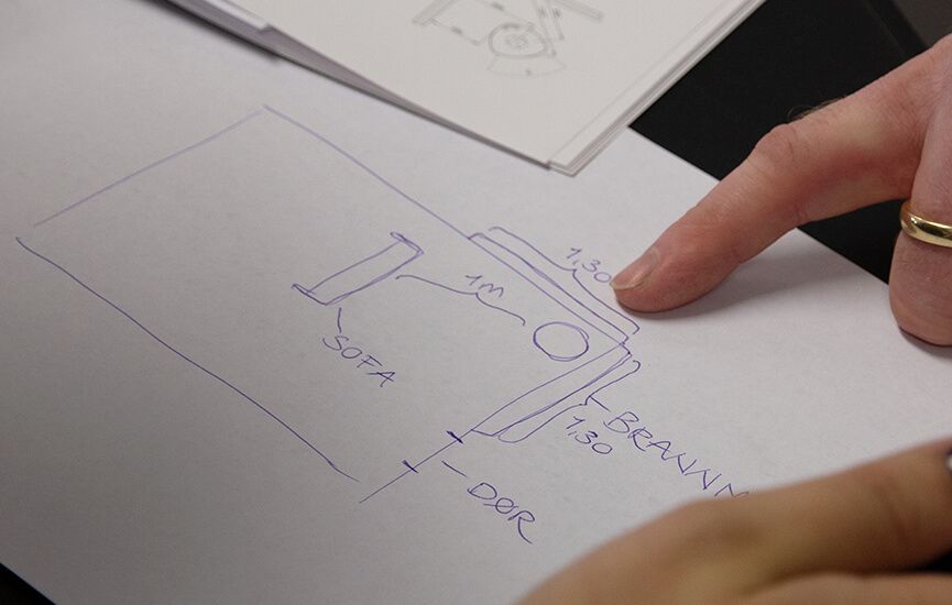 Hånd som peker på enkel skisse av rom med planlagt plassering av peis, sofa og avstander