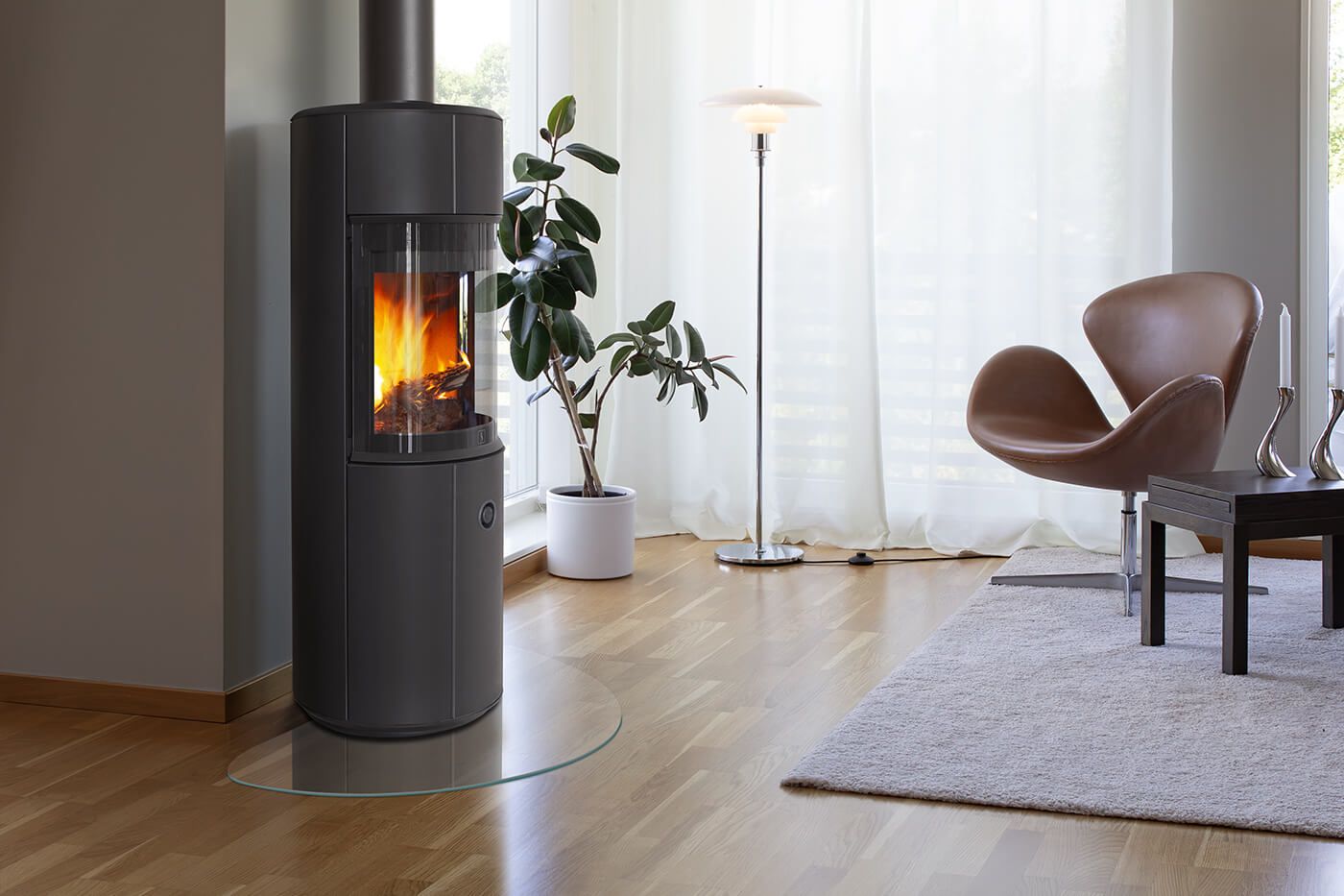 Frittstående vedovn med synlige flammer plassert i stue, med lenestol, gulvlampe og plante i bakgrunnen