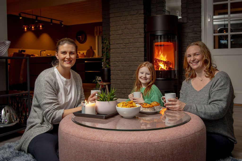 Familie sitter rundt salongbord i stue med frittstående vedovn og synlige flammer i bakgrunnen