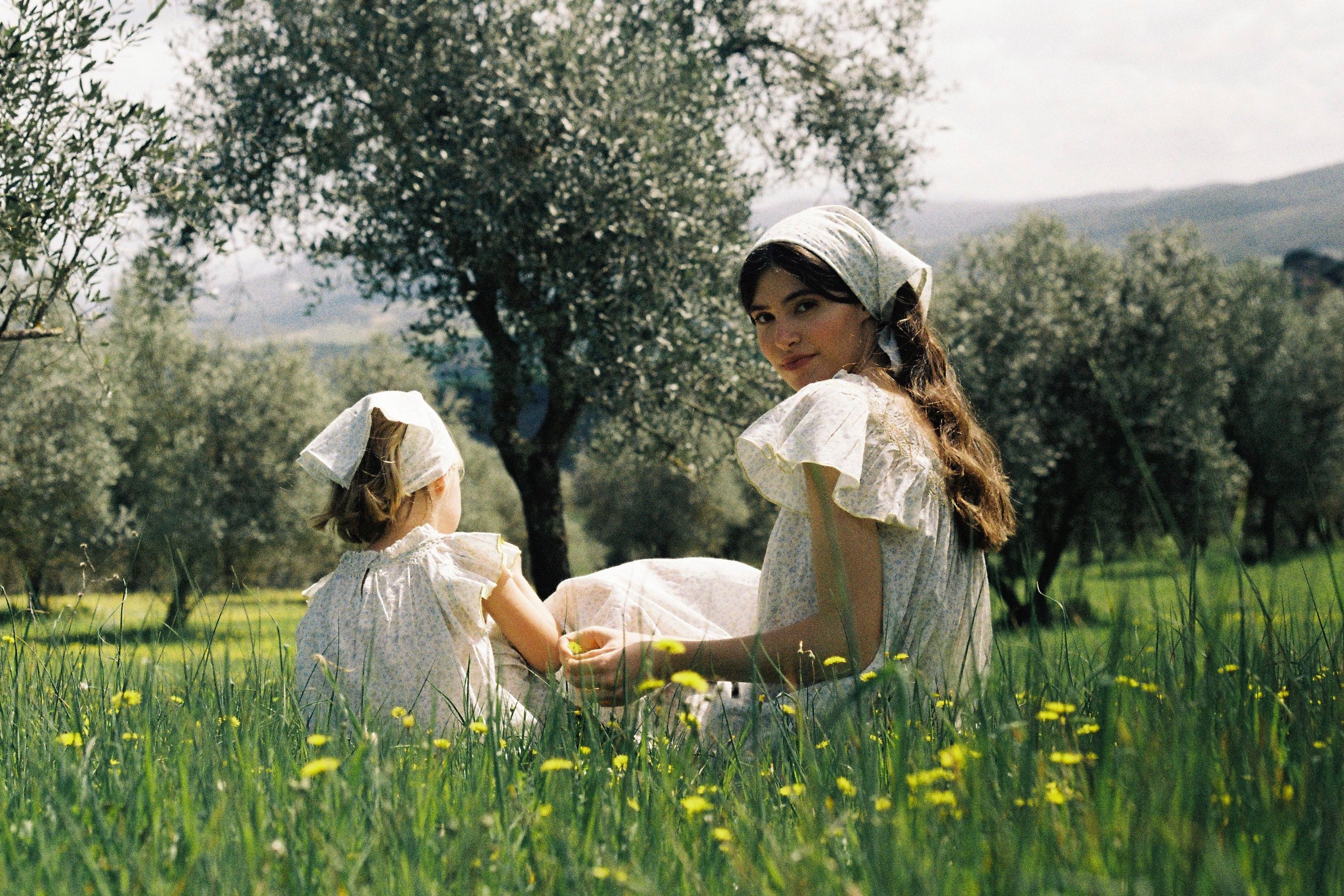 Woman and child sitting in a meadow wearing night dresses surrounded by trees and flowers